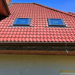Czerwony dach z dwoma oknami dachowymi marki Fakro w niebieski dzień. Widoczne rynny i część elewacji budynku.
