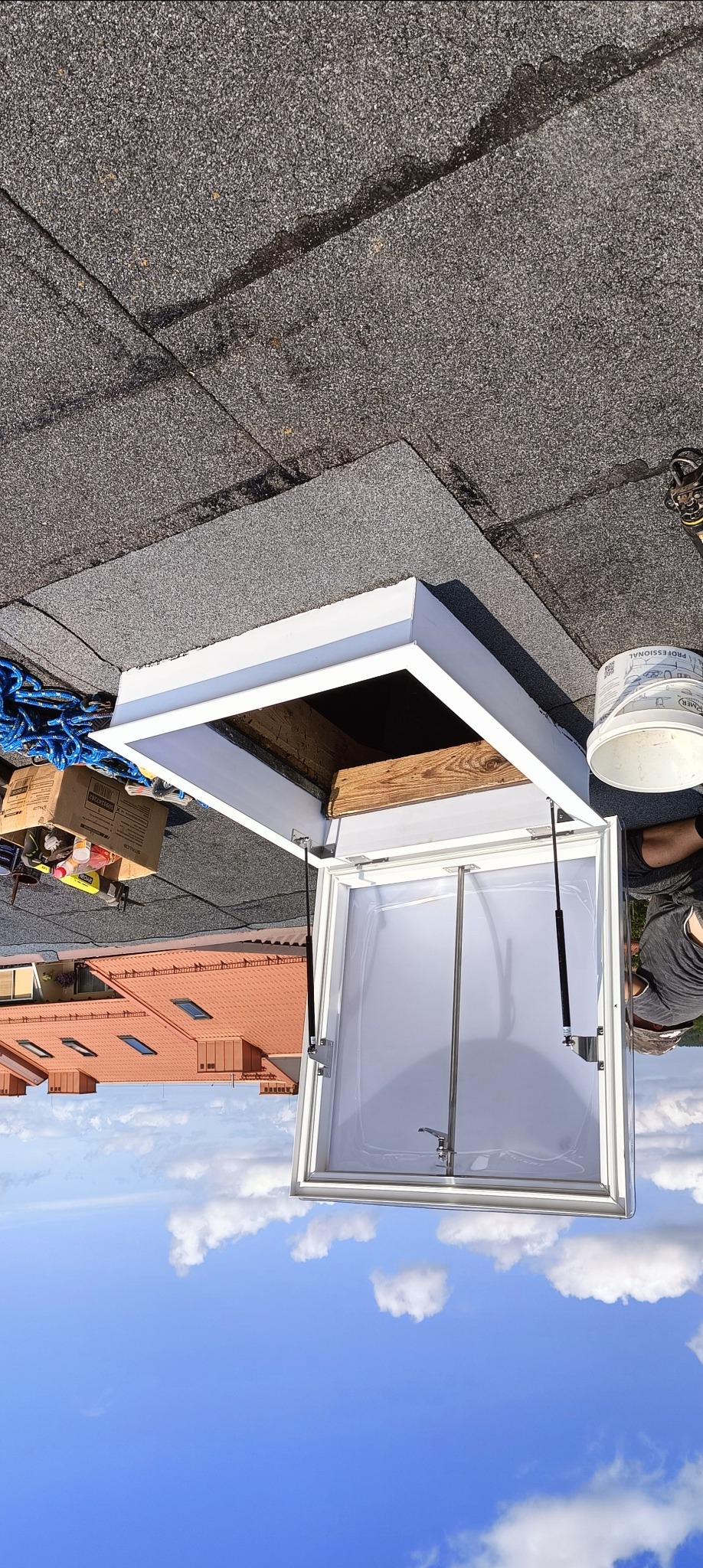 Otwarty, biały wyłaz dachowy na bitumicznym dachu. Widoczne narzędzia i wiadro. W tle budynek mieszkalny i błękitne niebo. Ujęcie z dołu, obrócone o 180 stopni.