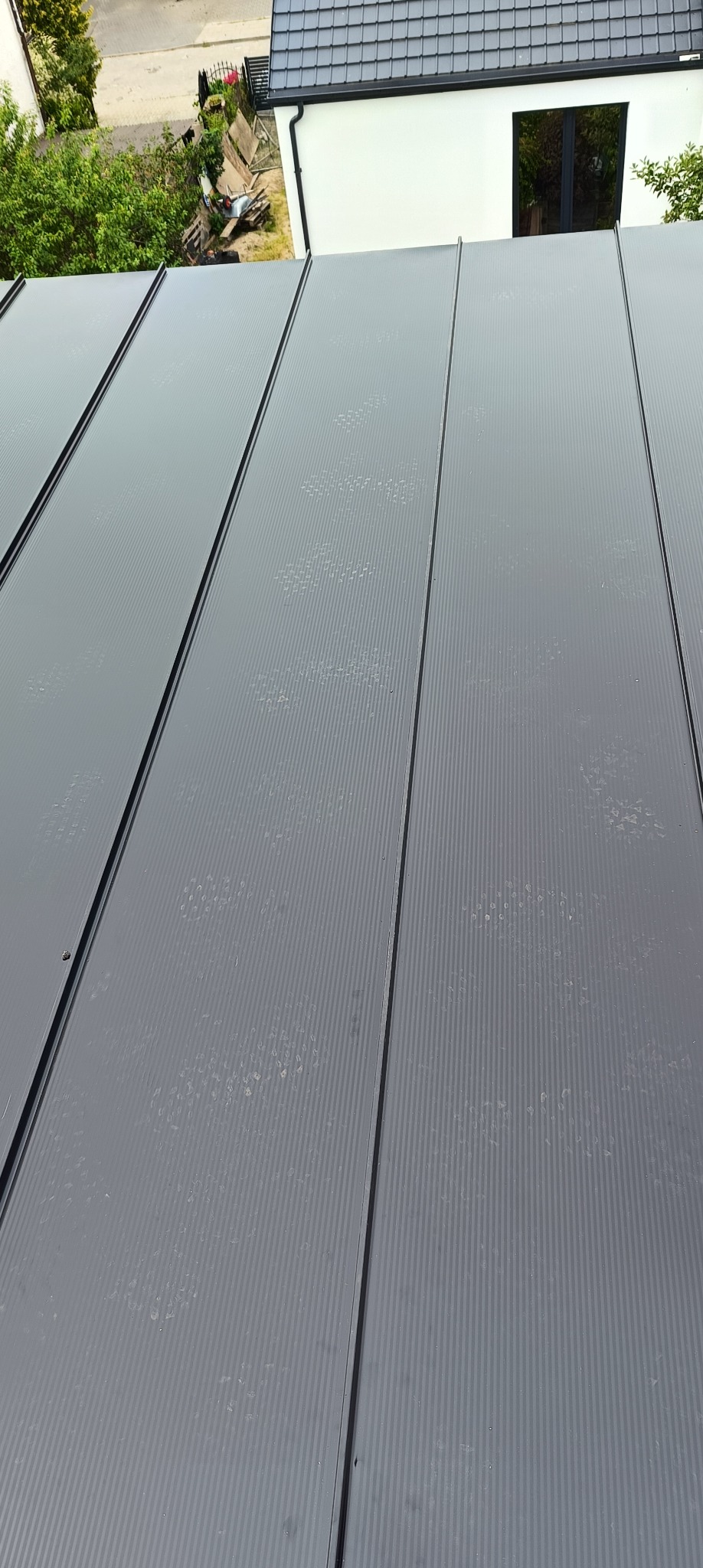 Nowoczesny, grafitowy dach z blachy trapezowej, widok z góry, na tle fragmentu budynku z czarną ramą okienną i zieleni. Widoczne ślady po deszczu na powierzchni.