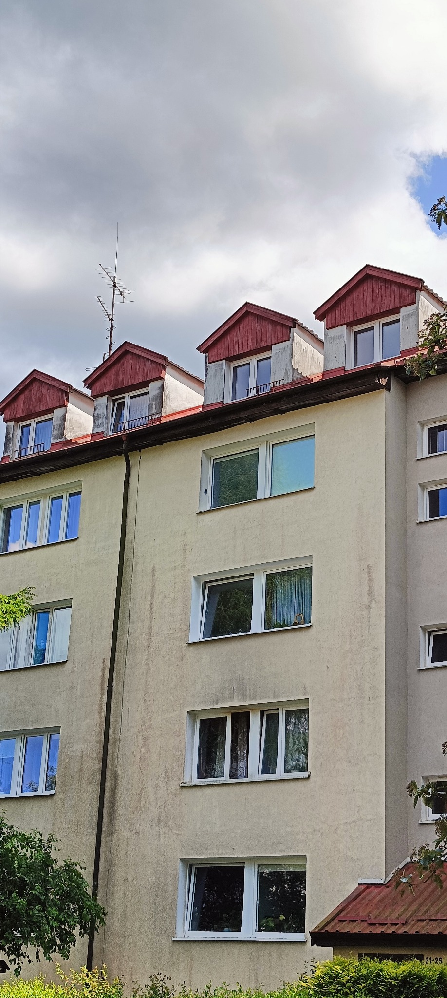 Elewacja bloku z lukarnami o czerwonych dachach i anteną na dachu. Widoczne okna z odbiciami zieleni i fragmenty roślinności. Budynek o jasnej, lekko zabrudzonej elewacji.