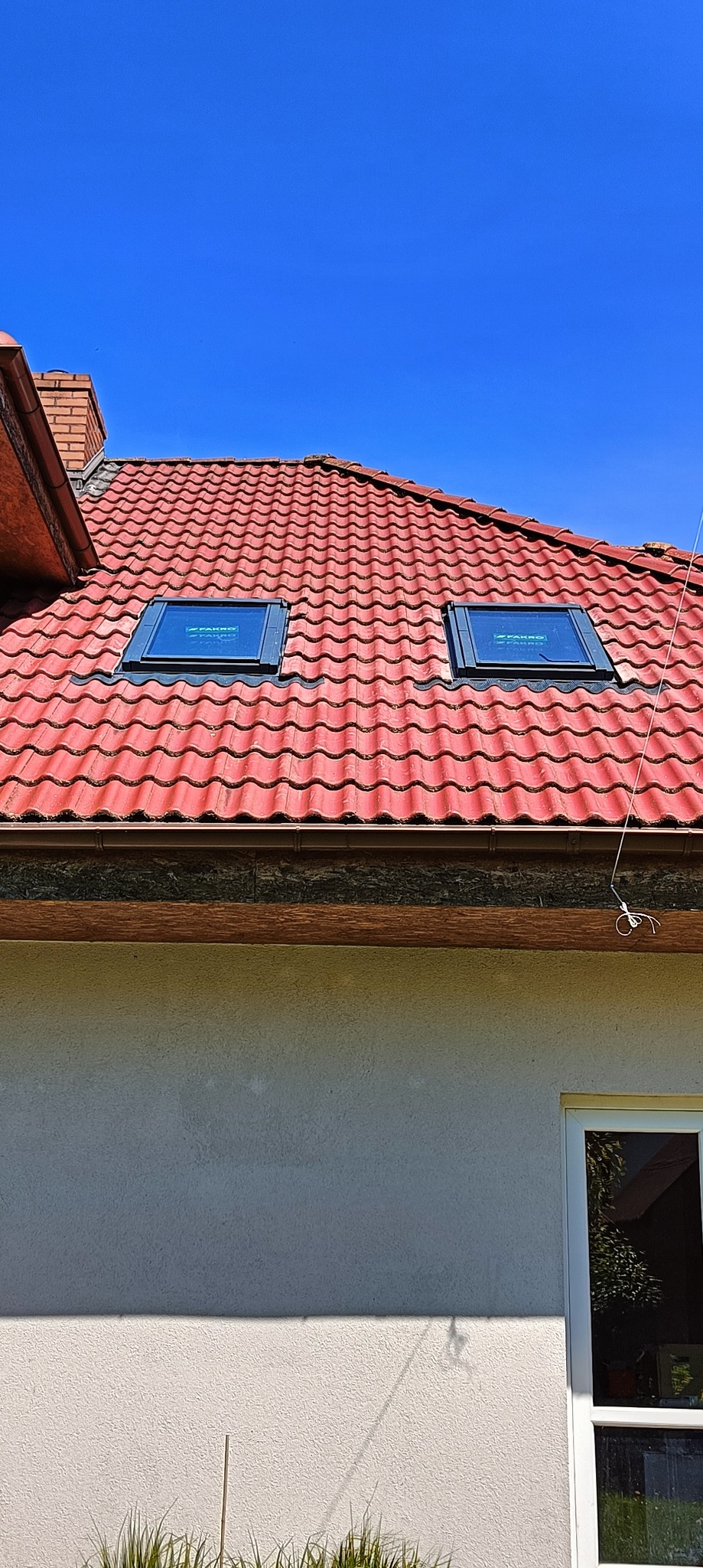 Czerwony dach z dwoma oknami dachowymi marki Fakro w niebieski dzień. Widoczne rynny i część elewacji budynku.