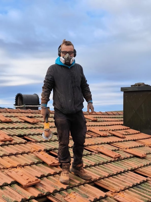 Pracownik w masce i słuchawkach ochronnych, stojący na dachu z dachówki ceramicznej z szlifierką kątową w dłoni, widoczny komin w tle.