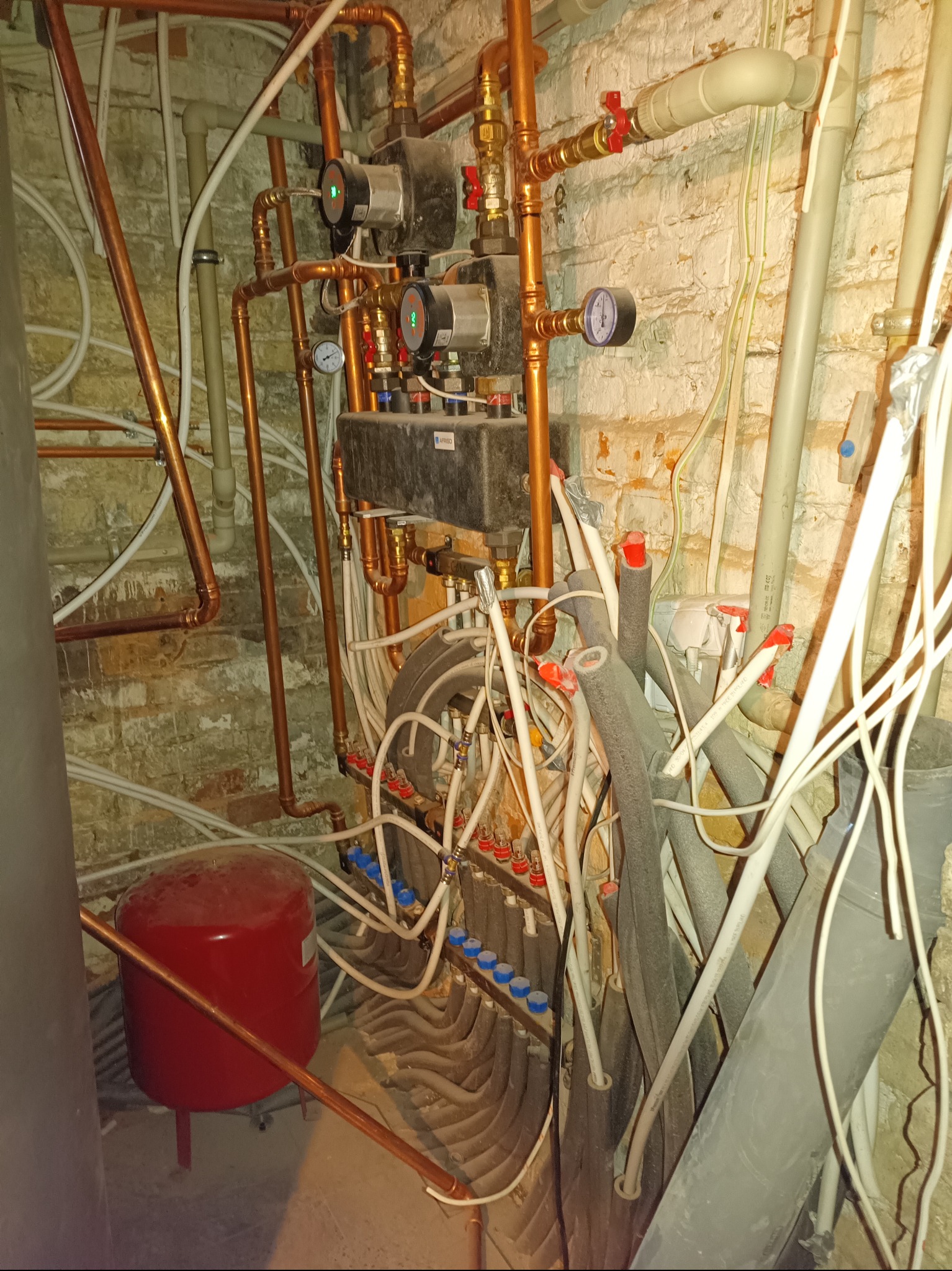 Złożona instalacja hydrauliczna z rur miedzianych, zaworów, manometrów i rozdzielacza, widoczna na tle ściany z cegły pomalowanej na biało.