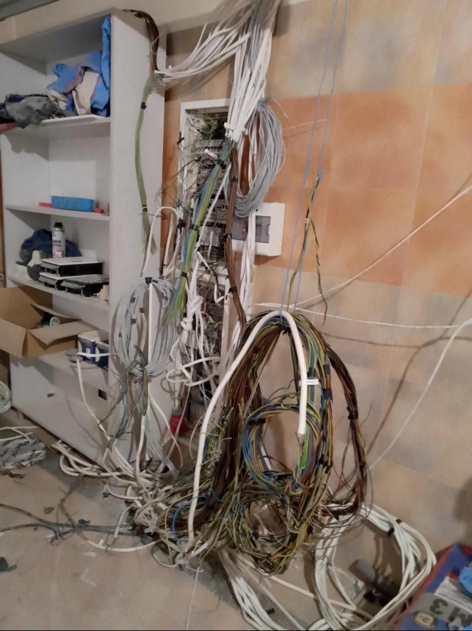 Skomplikowana instalacja elektryczna w trakcie montażu, widoczne liczne przewody, puszka rozdzielcza i fragment regału z narzędziami.