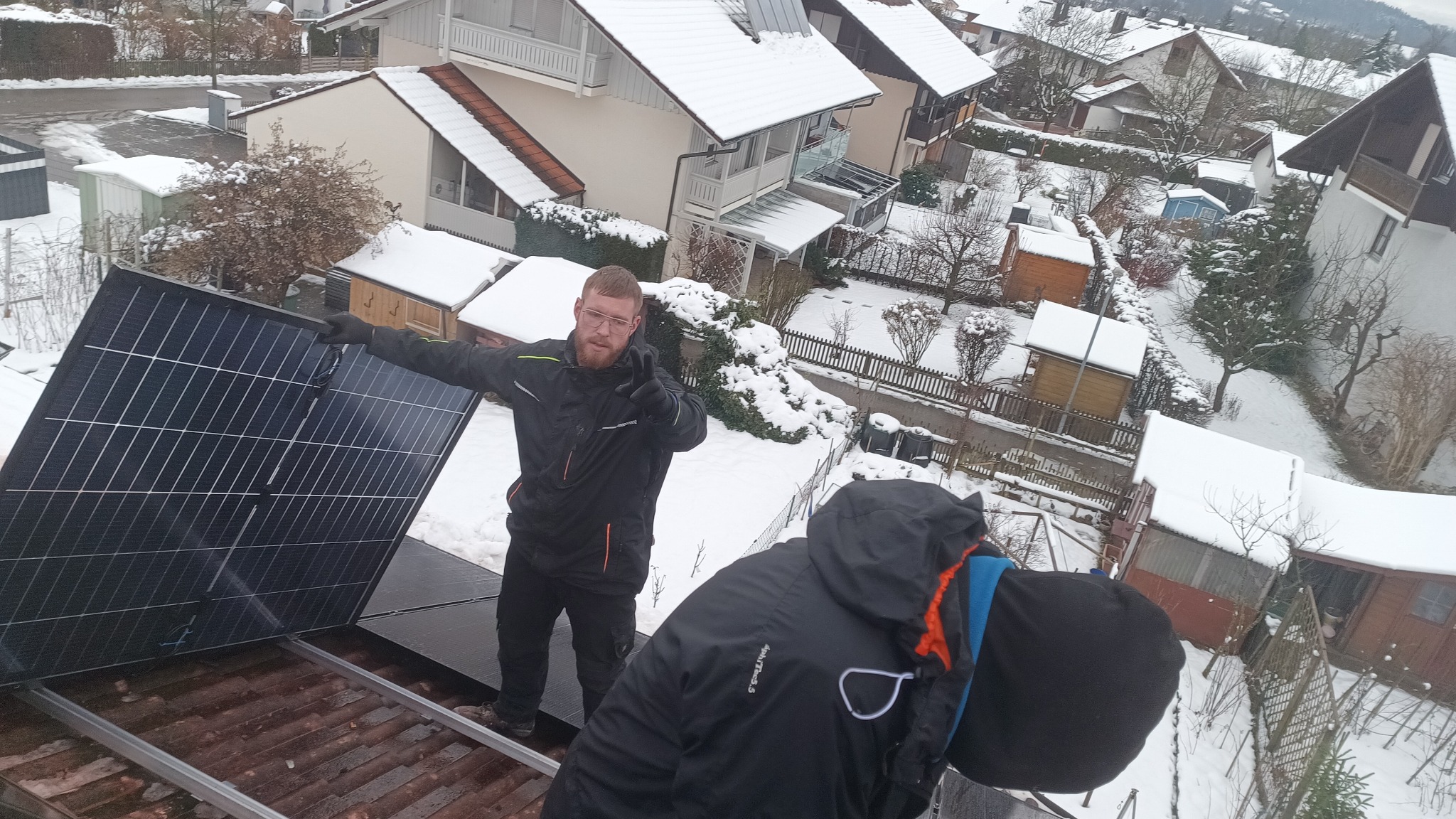 Montaż paneli słonecznych na dachu domu pokrytym śniegiem, widok z góry, instalator z brodą w czarnej kurtce pokazuje dwa palce, w tle zabudowa jednorodzinna.