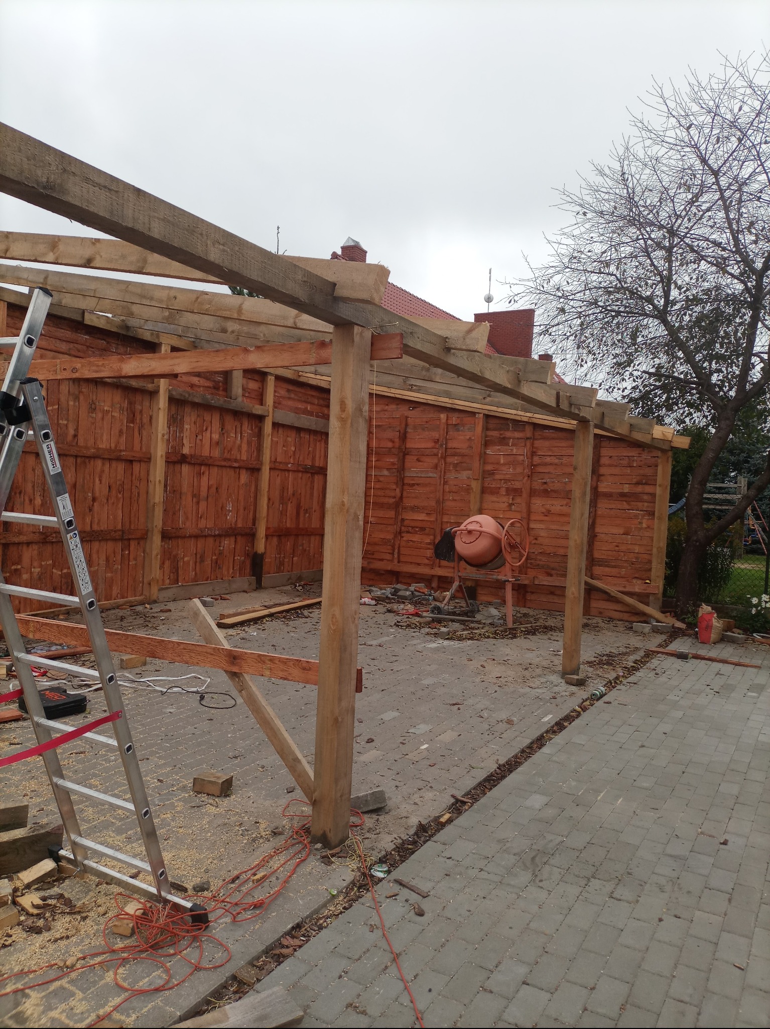 Budowa drewnianej konstrukcji z widocznymi deskami, słupami i elementami dachu, drabina oparta o ścianę, betoniarka w tle, teren budowy z resztkami materiałów.