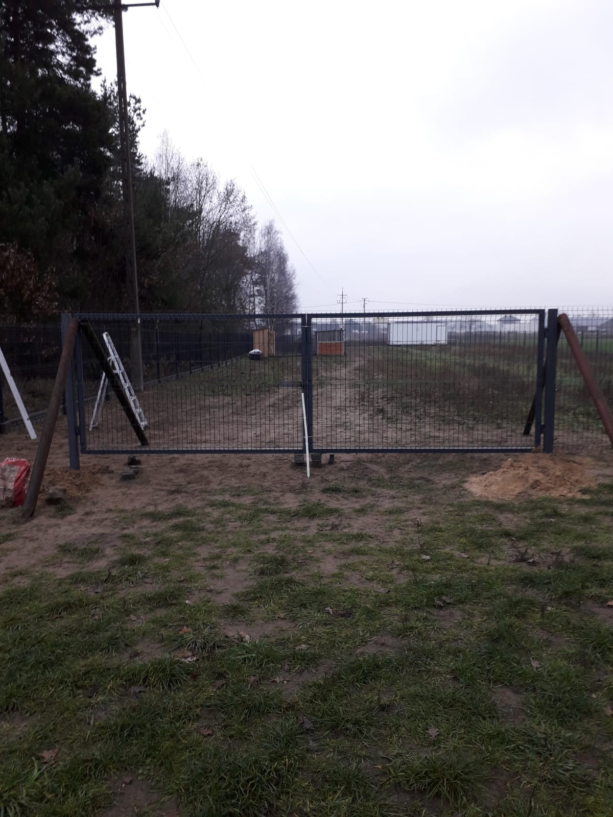 Nowo zamontowana, dwuskrzydłowa, metalowa brama ogrodzeniowa na posesji; widoczne elementy montażowe, drabina oparta o słupek oraz worki z zaprawą. Tło stanowią drzewa i budynki gospodarcze.