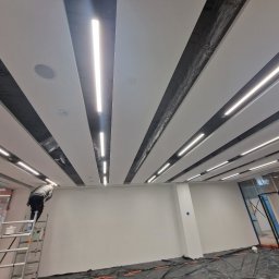 WK CONSTRUCTION SPÓŁKA Z OGRANICZONĄ ODPOWIEDZIALNOŚCIĄ - Pracownik na drabinie maluje sufit z podłużnymi panelami i oświetleniem LED w nowoczesnym biurze, folia ochronna na podłodze.