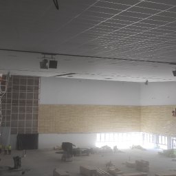 WK CONSTRUCTION SPÓŁKA Z OGRANICZONĄ ODPOWIEDZIALNOŚCIĄ - Wnętrze dużego pomieszczenia w trakcie prac wykończeniowych; widoczne rusztowania z drewnianą konstrukcją, podnośniki koszowe z pracownikami, ściany z częściowo położoną izolacją oraz sufit...