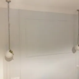 Szara ściana z dekoracyjnymi panelami i dwoma wiszącymi lampami kulistymi z mosiężnymi elementami, widoczne gniazdko elektryczne.