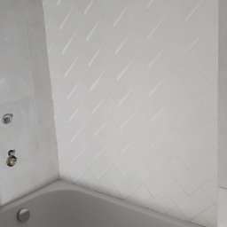 WK CONSTRUCTION SPÓŁKA Z OGRANICZONĄ ODPOWIEDZIALNOŚCIĄ - Biała wanna z widoczną ścianą wyłożoną płytkami w jodełkę, armatura w trakcie montażu.