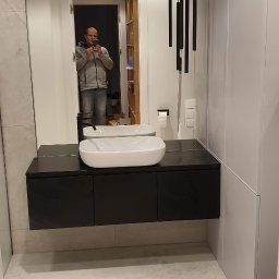 WK CONSTRUCTION SPÓŁKA Z OGRANICZONĄ ODPOWIEDZIALNOŚCIĄ - Nowoczesna łazienka z białym umywalką nablatową, czarną szafką pod umywalką, wiszącą toaletą i podłużnymi lampami wiszącymi, widok odbity w dużym lustrze z mężczyzną robiącym zdjęcie.