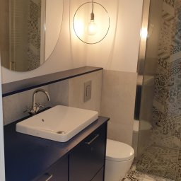 WK CONSTRUCTION SPÓŁKA Z OGRANICZONĄ ODPOWIEDZIALNOŚCIĄ - Nowoczesna łazienka z okrągłym lustrem, wiszącą lampą z żarówką Edisona, umywalką nablatową na ciemnoniebieskiej szafce z szufladami oraz kabiną prysznicową z wzorzystymi płytkami.