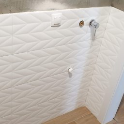 WK CONSTRUCTION SPÓŁKA Z OGRANICZONĄ ODPOWIEDZIALNOŚCIĄ - Białe, trójwymiarowe płytki na ścianie w trakcie montażu, widoczne niedokończone wykończenie ściany, przygotowane przyłącza wodne i włącznik światła, drewnopodobna podłoga.