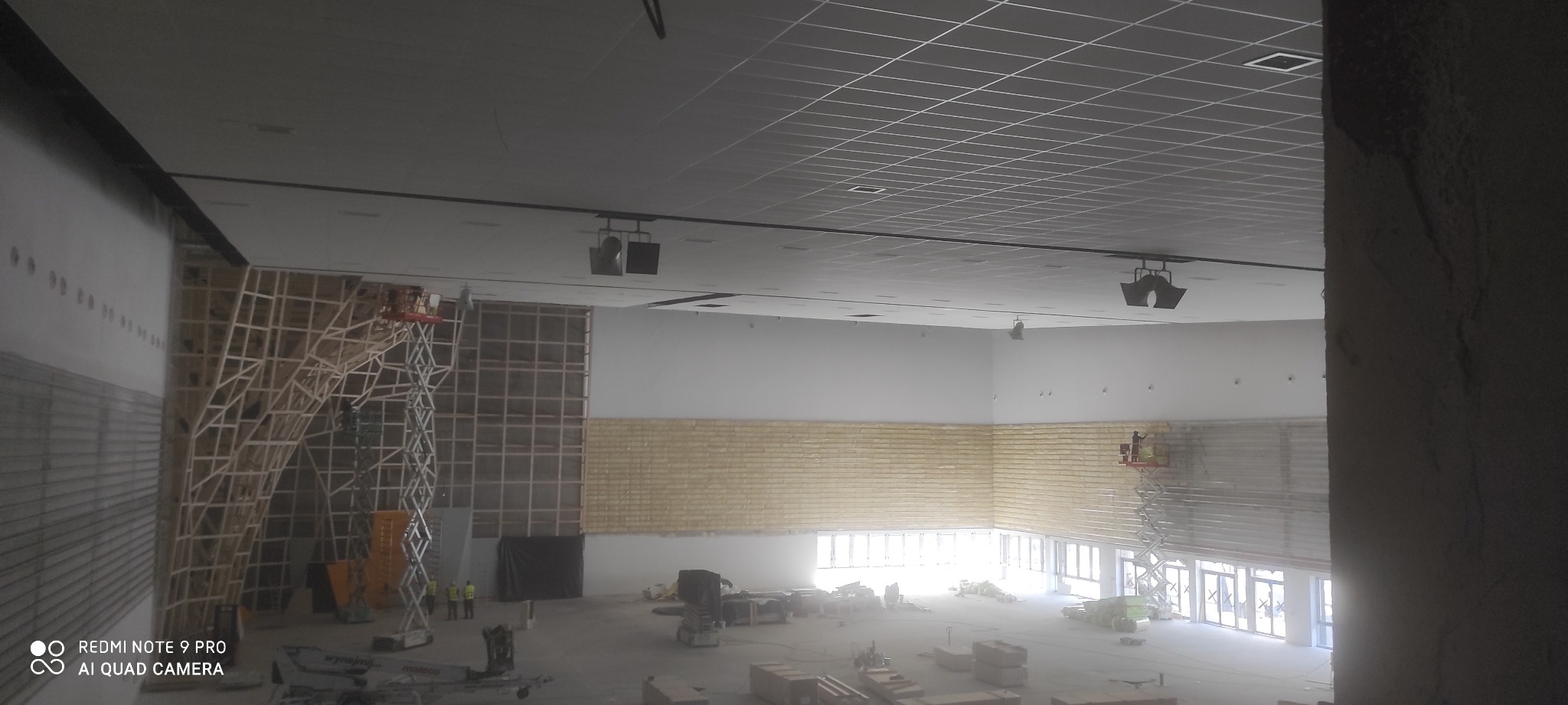 Wnętrze dużego pomieszczenia w trakcie prac wykończeniowych; widoczne rusztowania z drewnianą konstrukcją, podnośniki koszowe z pracownikami, ściany z częściowo położoną izolacją oraz sufit...