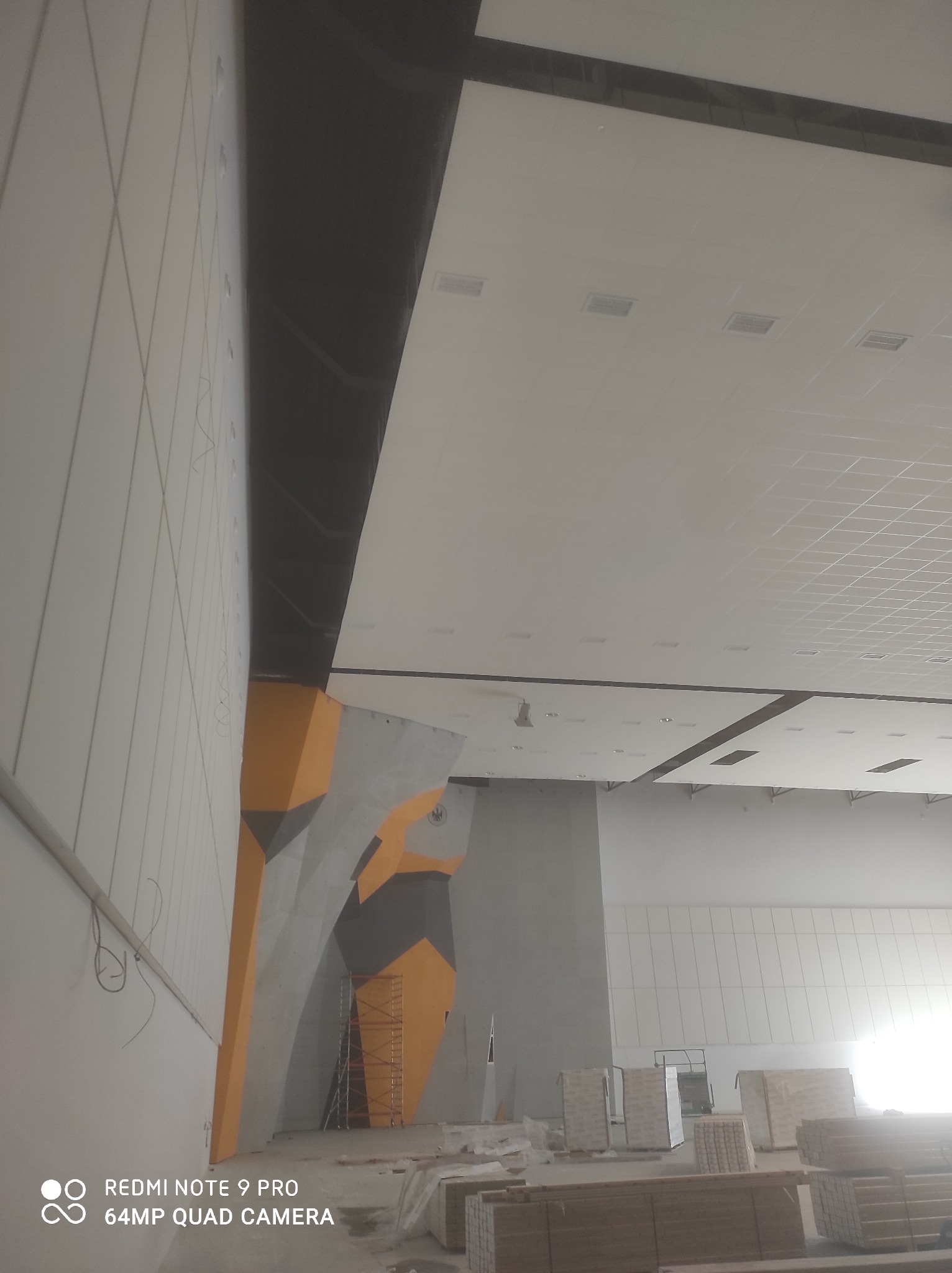 Montaż sufitu podwieszanego z widocznymi elementami konstrukcyjnymi i przygotowanymi materiałami, ściana z elementami dekoracyjnymi w kolorach szarym i pomarańczowym, oraz rusztowanie w tle.