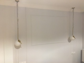 Szara ściana z dekoracyjnymi panelami i dwoma wiszącymi lampami kulistymi z mosiężnymi elementami, widoczne gniazdko elektryczne.