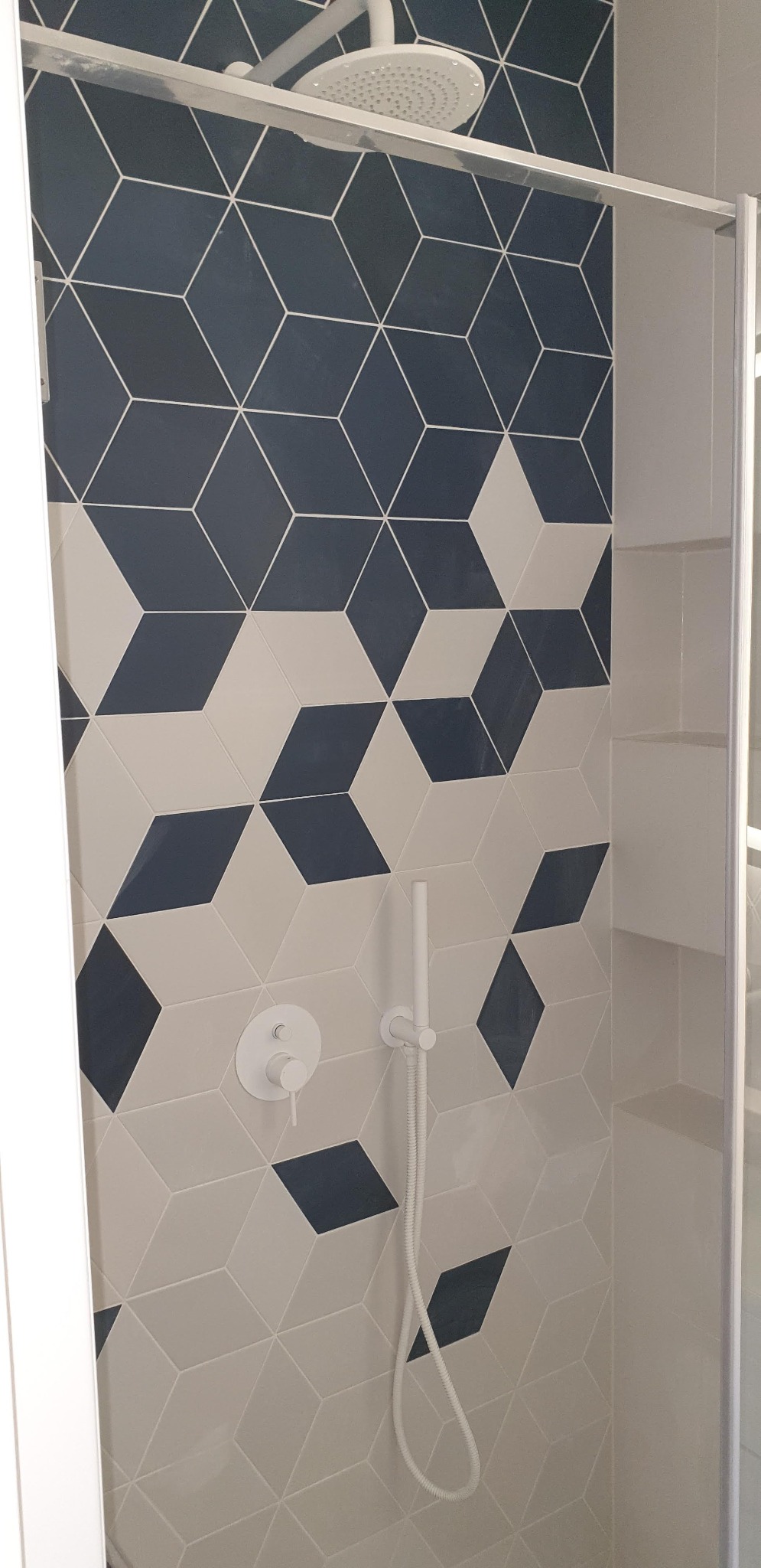 Kabina prysznicowa z geometrycznymi płytkami w odcieniach granatu i beżu, biała armatura i półki wnękowe.