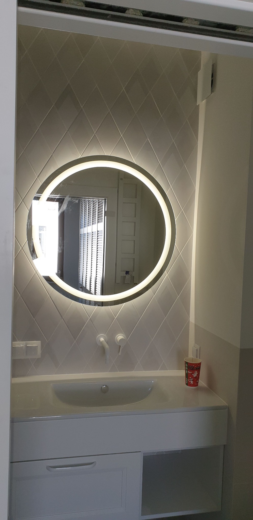 Nowoczesna łazienka z owalnym lustrem podświetlanym LED, białą umywalką nablatową i płytkami w romby na ścianie, widoczne białe baterie i kubek z motywem świątecznym.