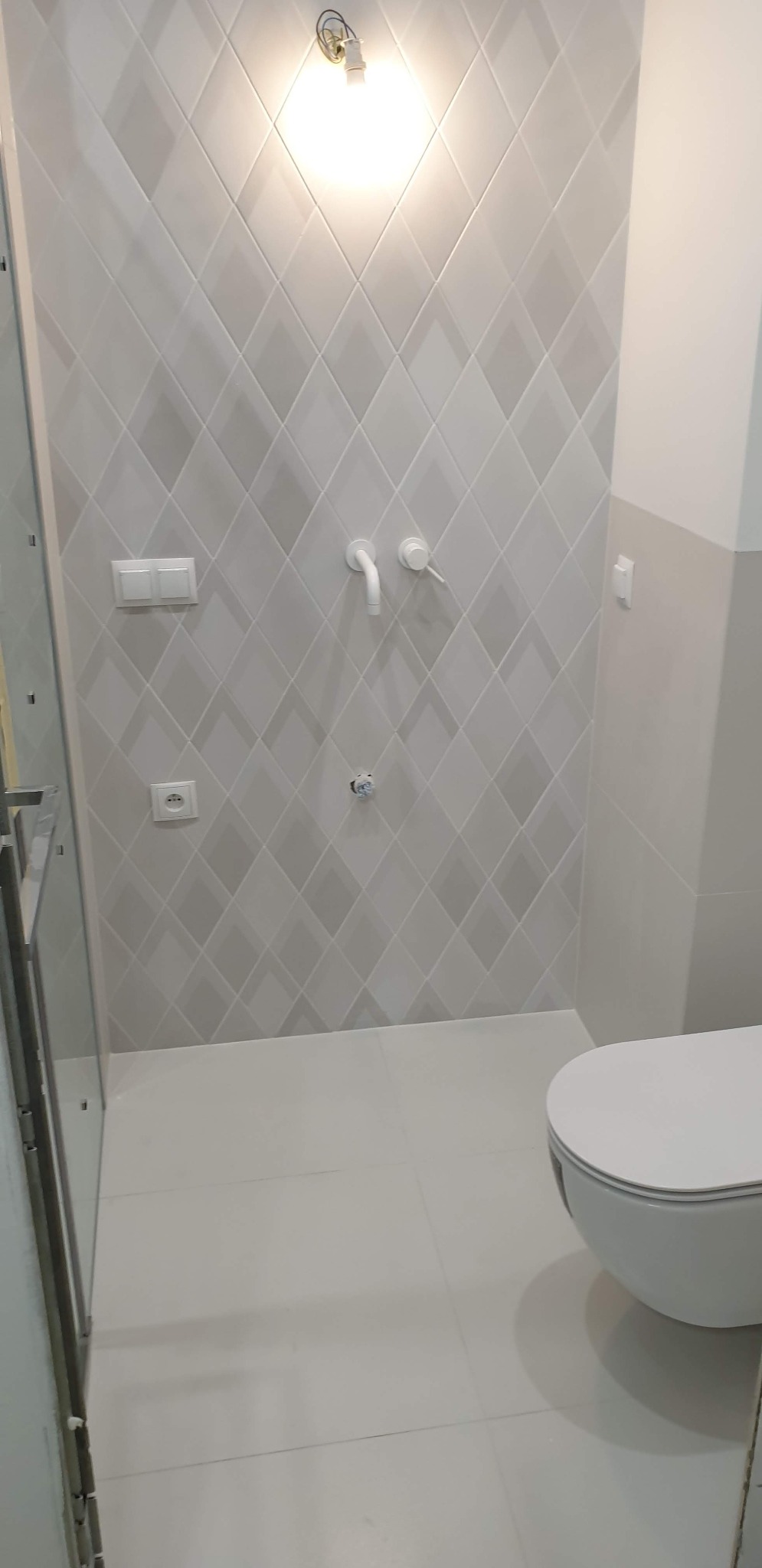 Jasnoszara łazienka z romboidalnymi płytkami na ścianach, białą armaturą i częściowo zamontowaną kabiną prysznicową po lewej stronie.