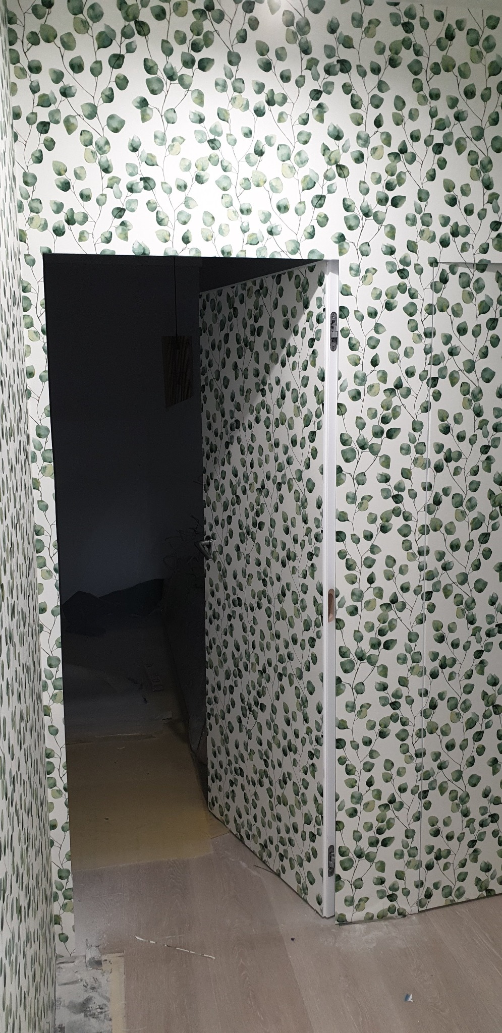 Ściana i drzwi oklejone tapetą z motywem eukaliptusa, widoczny fragment podłogi i ciemne wnętrze pomieszczenia za drzwiami.