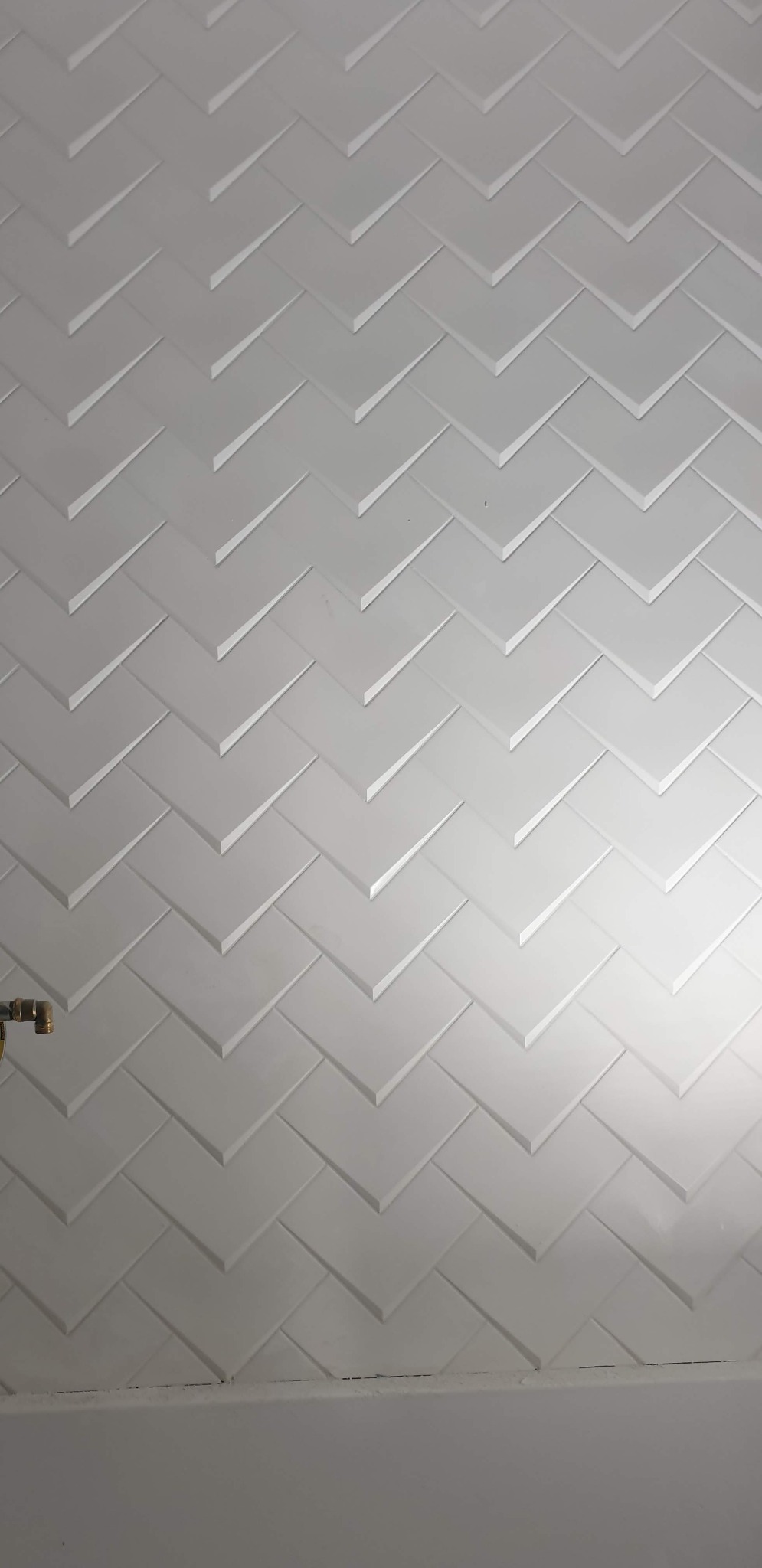 Biała ściana wykończona trójwymiarowymi płytkami w kształcie strzałek, ułożonych w jodełkę, z widocznym fragmentem metalowej rurki przy ścianie.