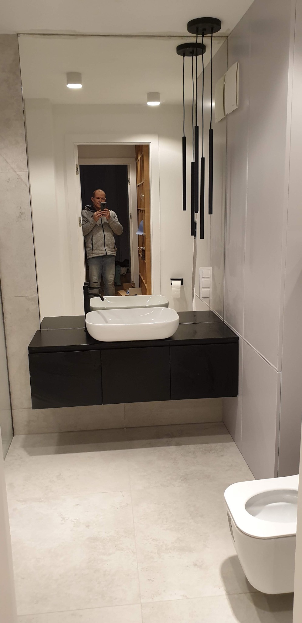 Nowoczesna łazienka z białym umywalką nablatową, czarną szafką pod umywalką, wiszącą toaletą i podłużnymi lampami wiszącymi, widok odbity w dużym lustrze z mężczyzną robiącym zdjęcie.