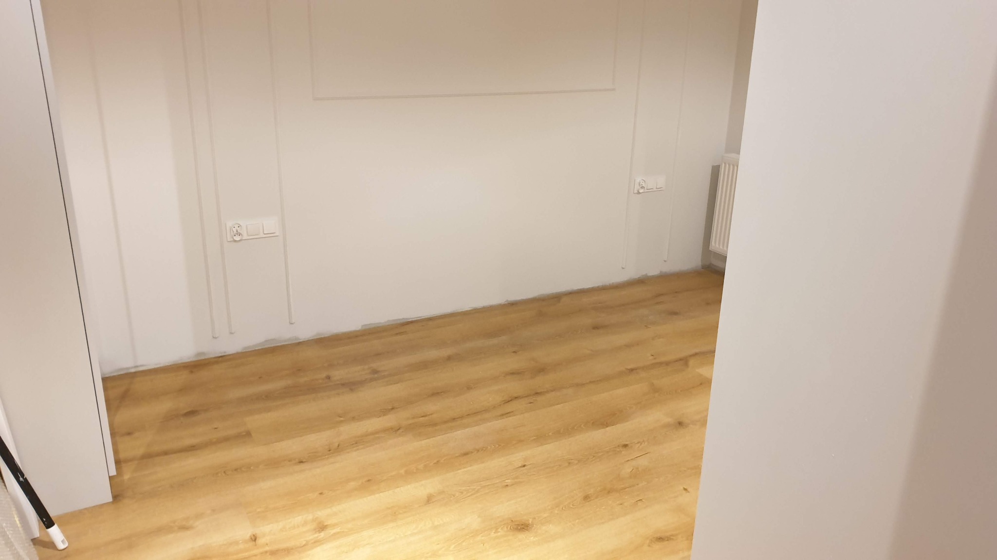 Jasne panele podłogowe w świeżo wykończonym pokoju z białymi ścianami ozdobionymi subtelnymi sztukateriami i widocznymi gniazdkami elektrycznymi.