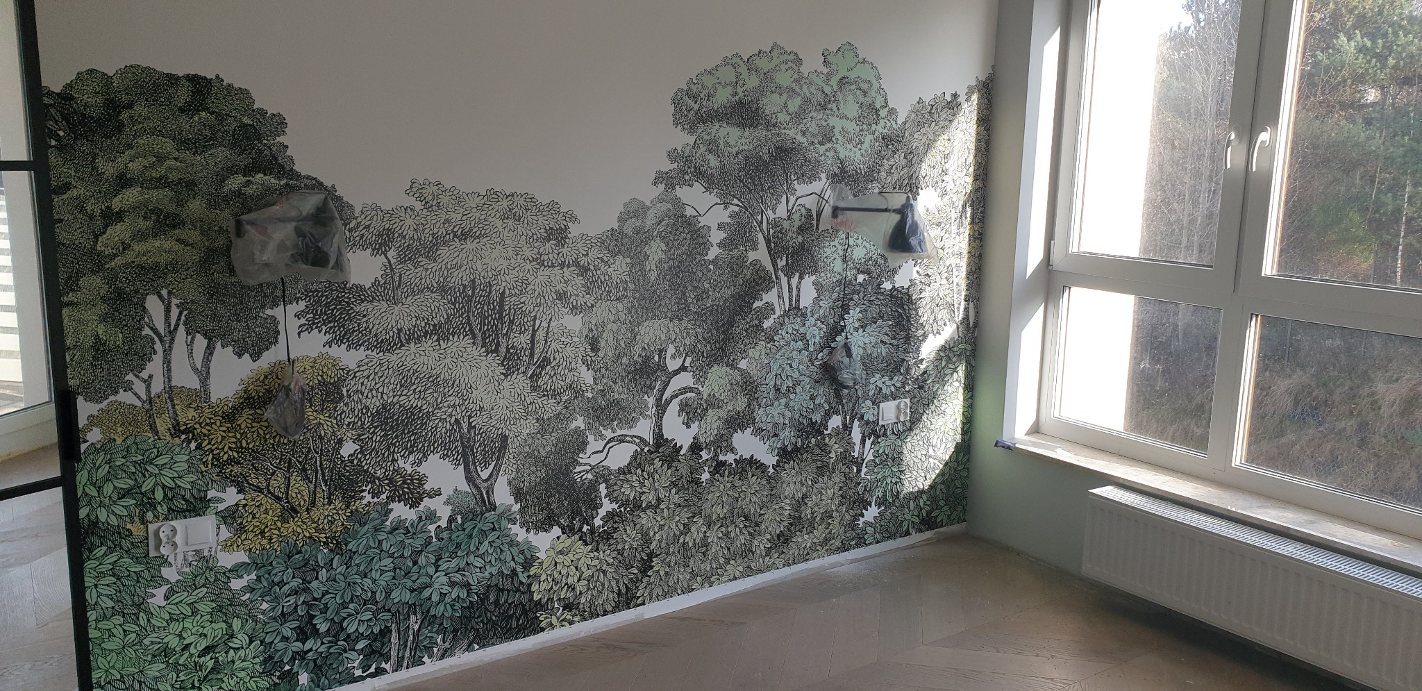 Ściana w Gdyni z fototapetą przedstawiającą motyw leśny, zabezpieczone gniazdka elektryczne folią malarską, parkiet ułożony w jodełkę.