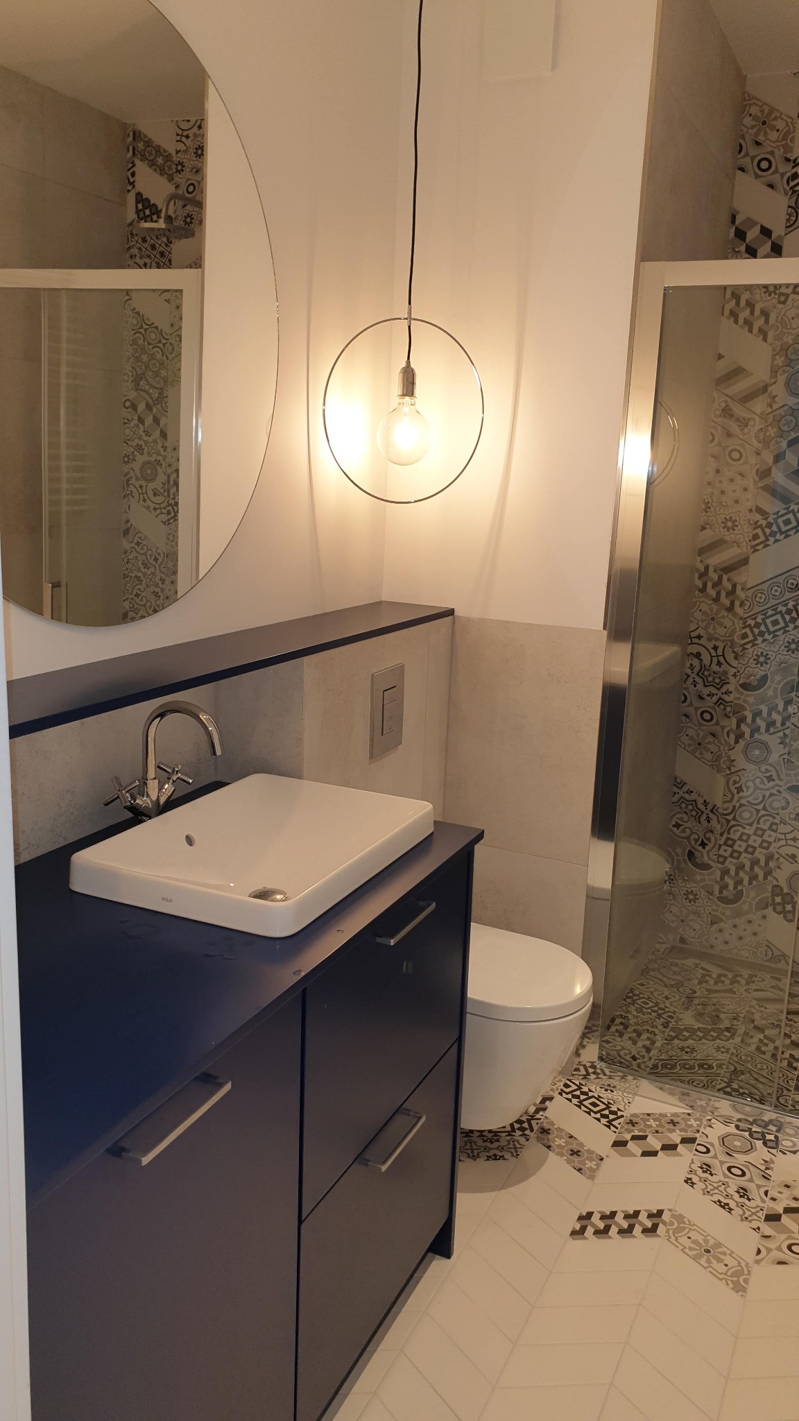 Nowoczesna łazienka z okrągłym lustrem, wiszącą lampą z żarówką Edisona, umywalką nablatową na ciemnoniebieskiej szafce z szufladami oraz kabiną prysznicową z wzorzystymi płytkami.