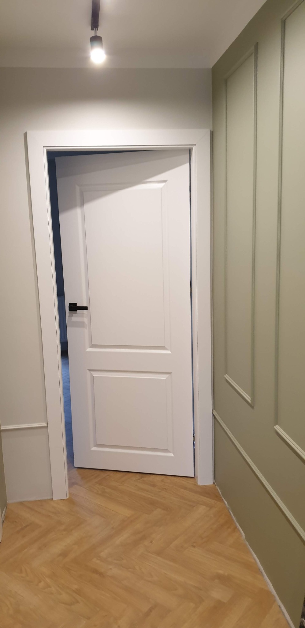 Białe drzwi wewnętrzne z czarną klamką, zamontowane w odnowionym wnętrzu z panelami podłogowymi ułożonymi w jodełkę i ścianami w odcieniach szarości i zieleni z dekoracyjnymi listwami.