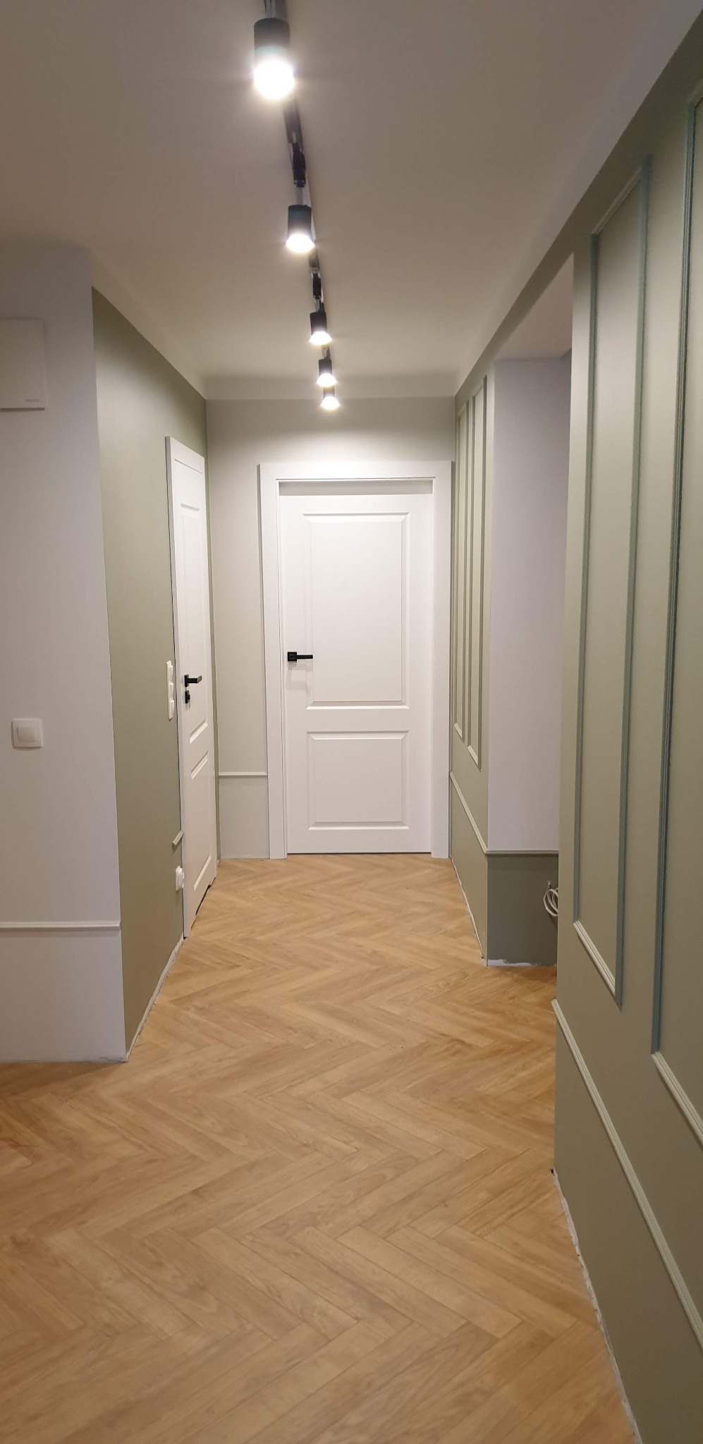 Korytarz po remoncie z parkietem ułożonym w jodełkę, ścianami w odcieniach bieli i oliwkowej zieleni, oświetleniem szynowym i sztukaterią.