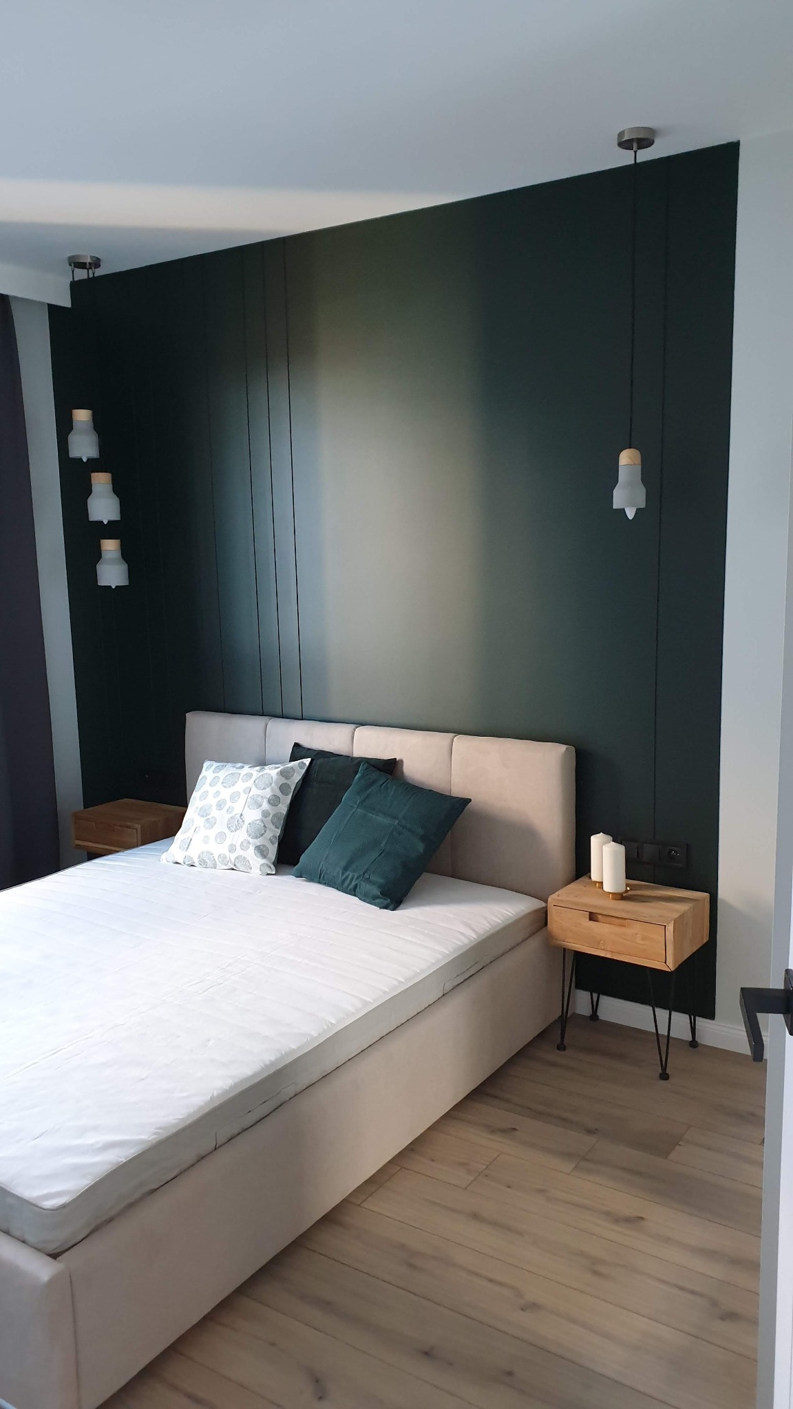 Nowoczesna sypialnia z ciemnozieloną ścianą z pionowymi panelami, beżowym łóżkiem i drewnianymi szafkami nocnymi na metalowych nóżkach, minimalistyczne lampy wiszące i ścienne.