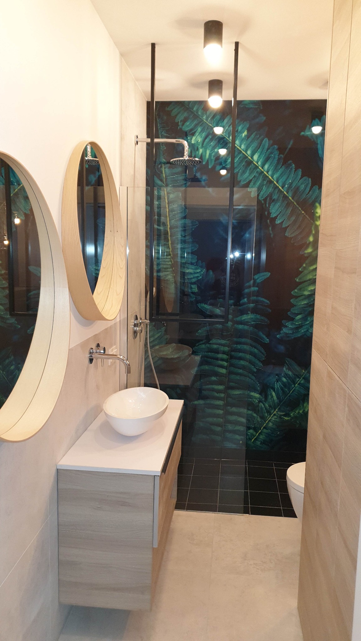 Nowoczesna łazienka z owalnymi lustrami w drewnianych ramach, białą umywalką nablatową i kabiną prysznicową z motywem tropikalnych liści na ścianie.