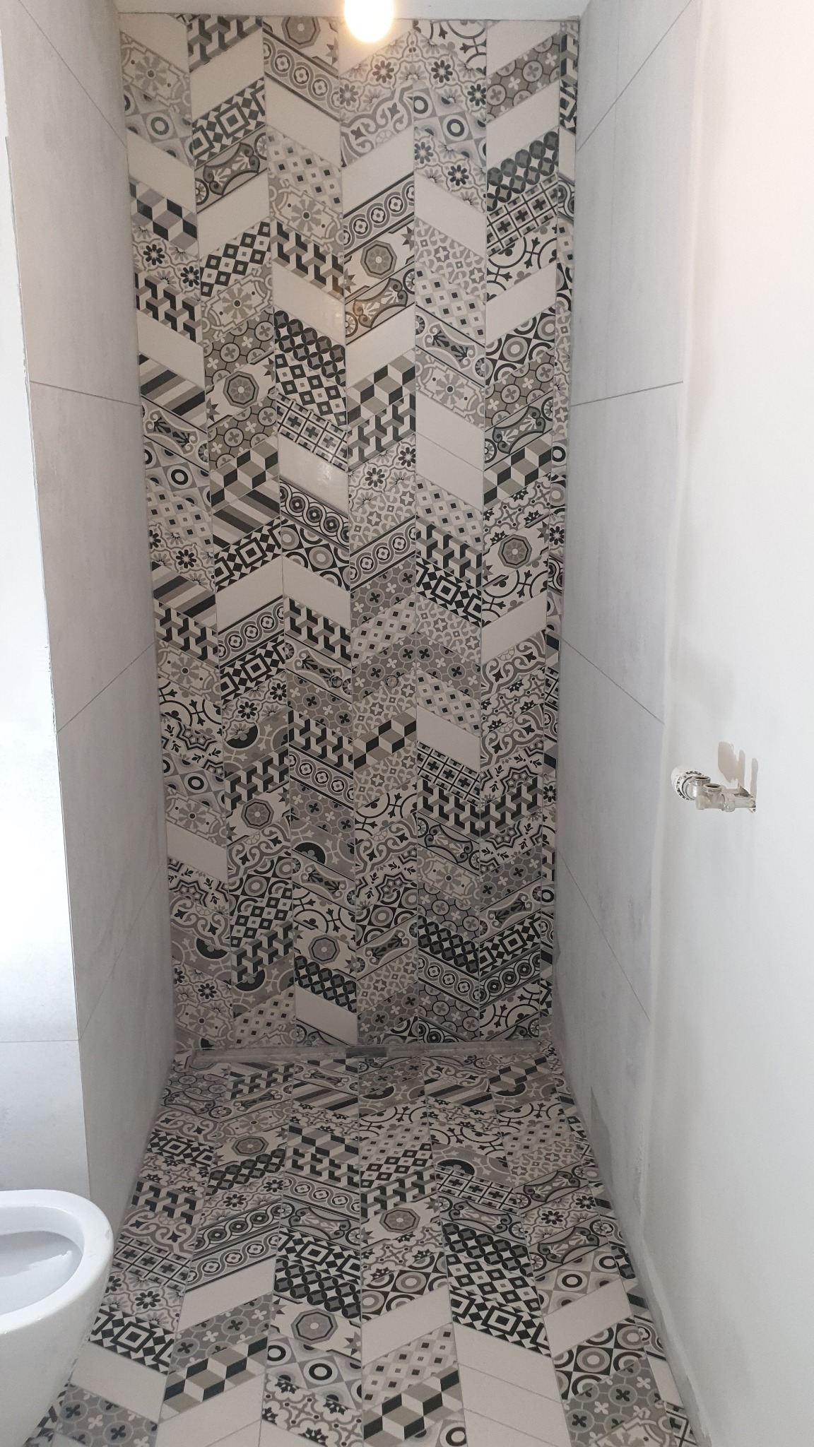 Wąska łazienka z wzorzystymi, czarno-białymi płytkami ułożonymi w jodełkę na ścianie i podłodze, widoczny fragment białej toalety i surowa ściana z instalacją wodną.