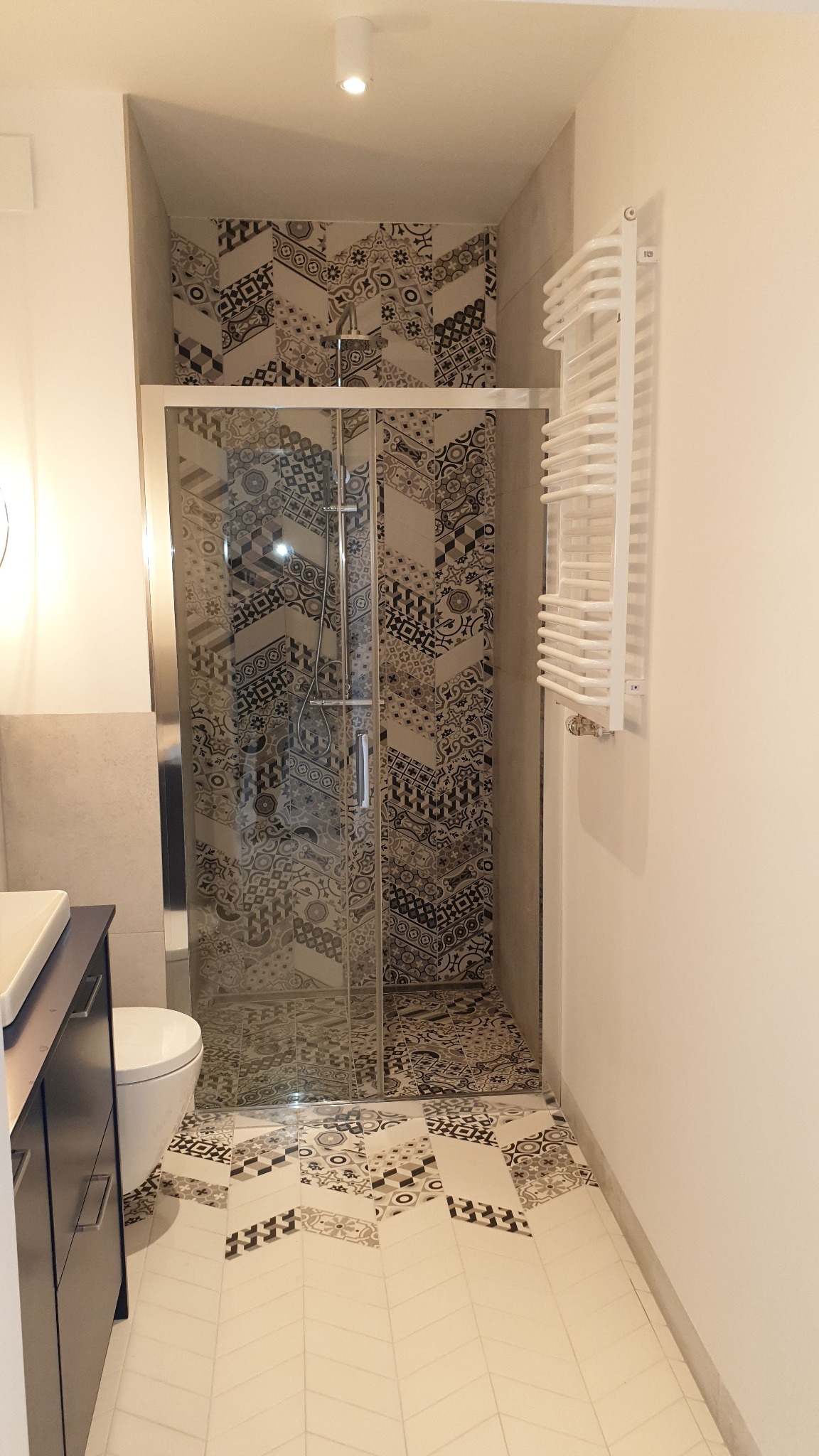 Nowoczesna łazienka z kabiną prysznicową wykończoną wzorzystymi płytkami, kontrastującymi z jasną podłogą ułożoną w jodełkę i białym grzejnikiem drabinkowym.