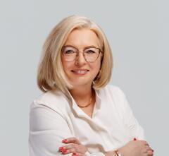 Portret uśmiechniętej blondynki w okularach i białej koszuli, z założonymi rękami, na jednolitym szarym tle. Profesjonalny wizerunek agenta nieruchomości.