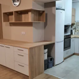 Jasna kuchnia z drewnianym blatem, białymi frontami szafek i dekoracyjnymi płytkami nad blatem roboczym; widoczna lodówka i szafki wiszące.