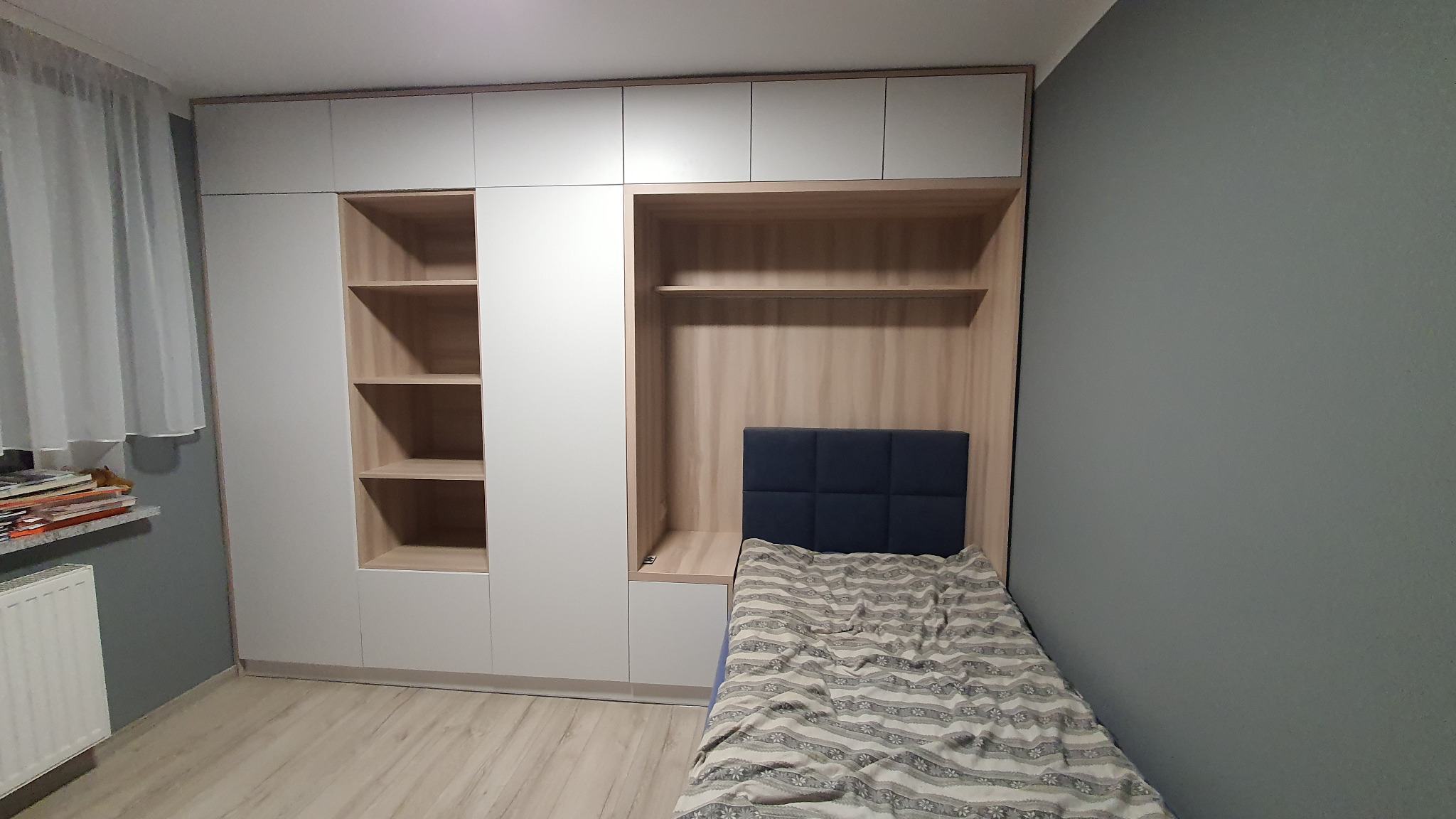 Biała szafa z półkami i wnęką na łóżko z tapicerowanym zagłówkiem w pokoju, widok z boku, na parapecie sterta książek.