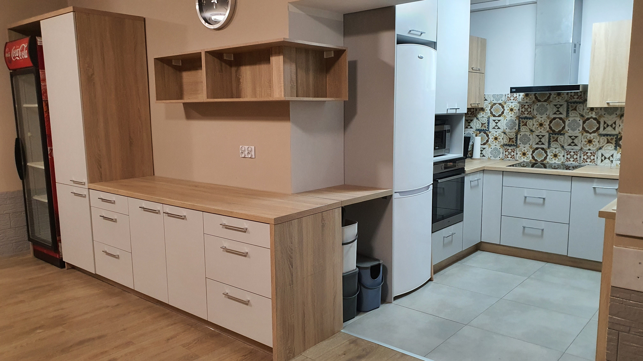 Jasna kuchnia z drewnianym blatem, białymi frontami szafek i dekoracyjnymi płytkami nad blatem roboczym; widoczna lodówka i szafki wiszące.