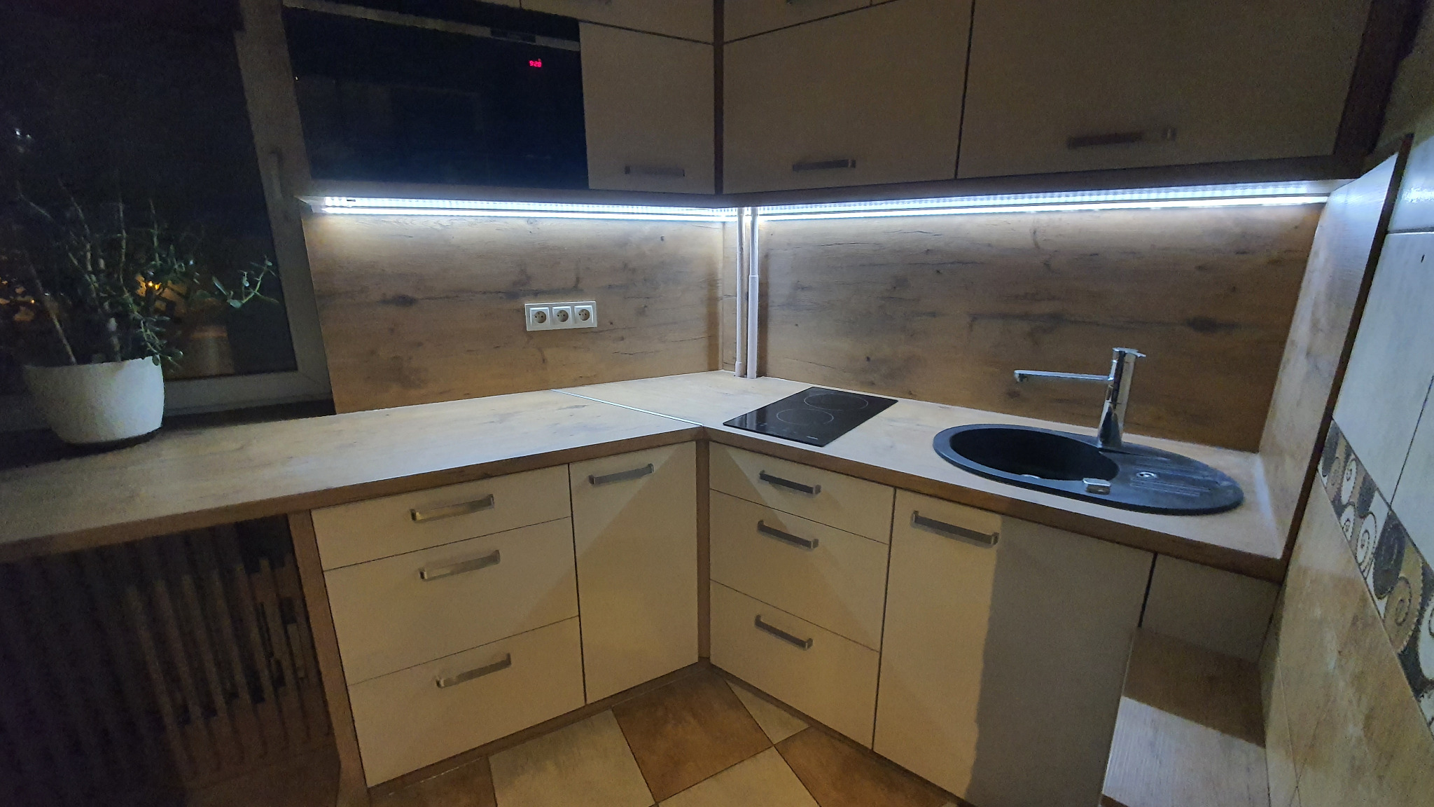 Nowoczesna kuchnia narożna z drewnianymi blatami, białymi frontami szafek i oświetleniem LED pod szafkami wiszącymi, widoczny zlew i płyta indukcyjna.