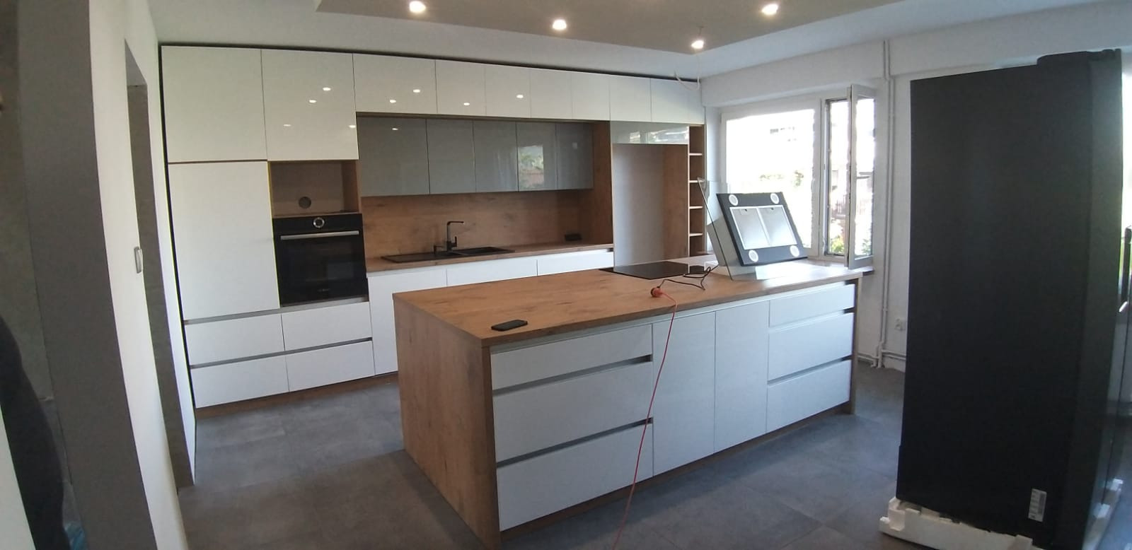 Nowoczesna kuchnia z białymi szafkami i wyspą z blatem w kolorze drewna, czarny piekarnik w zabudowie, szare szafki wiszące, widoczna lodówka w czarnym kolorze.