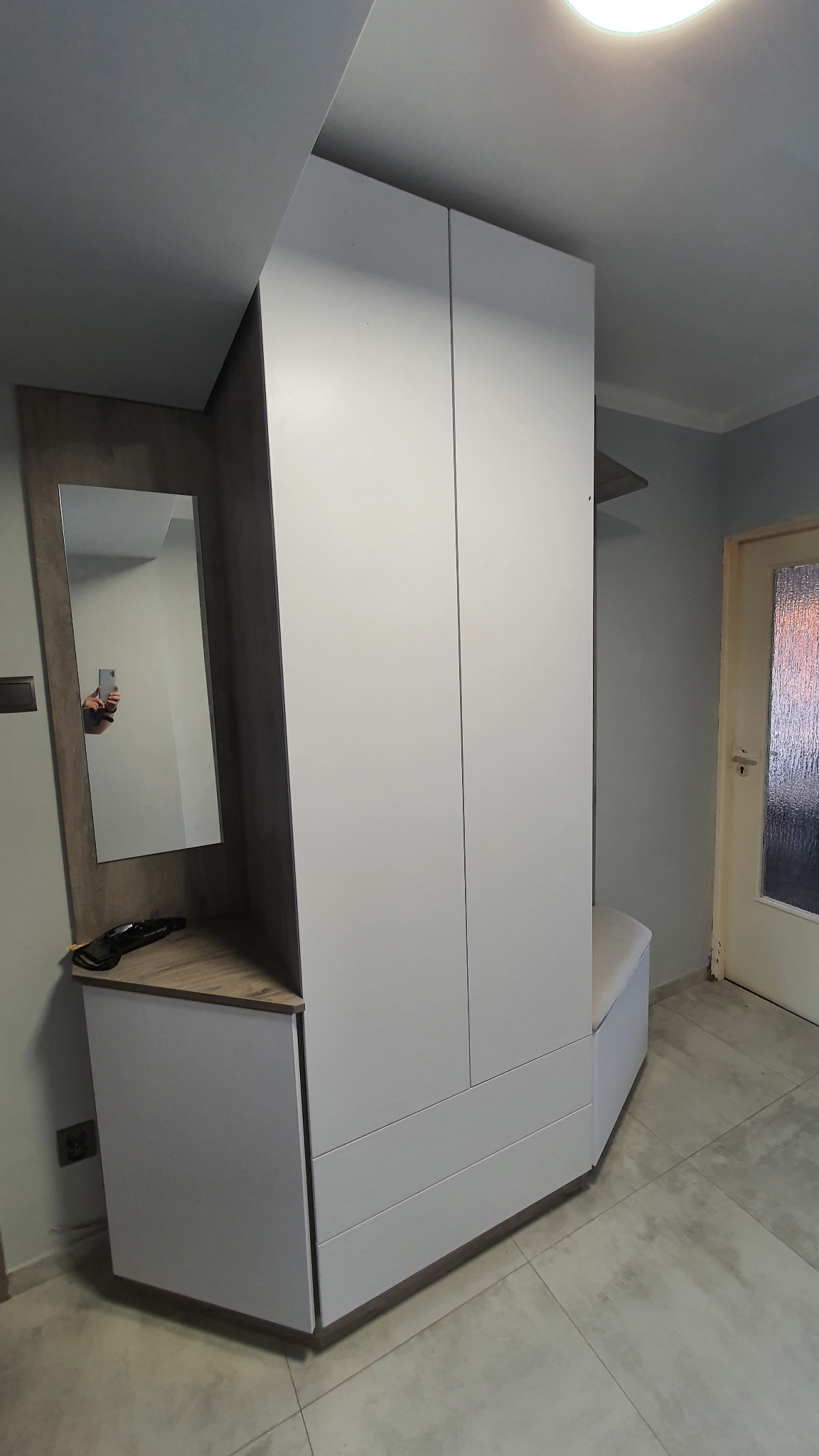Biała szafa na wymiar z siedziskiem i lustrem w przedpokoju, widoczne gniazdko elektryczne i odbicie fotografa w lustrze.
