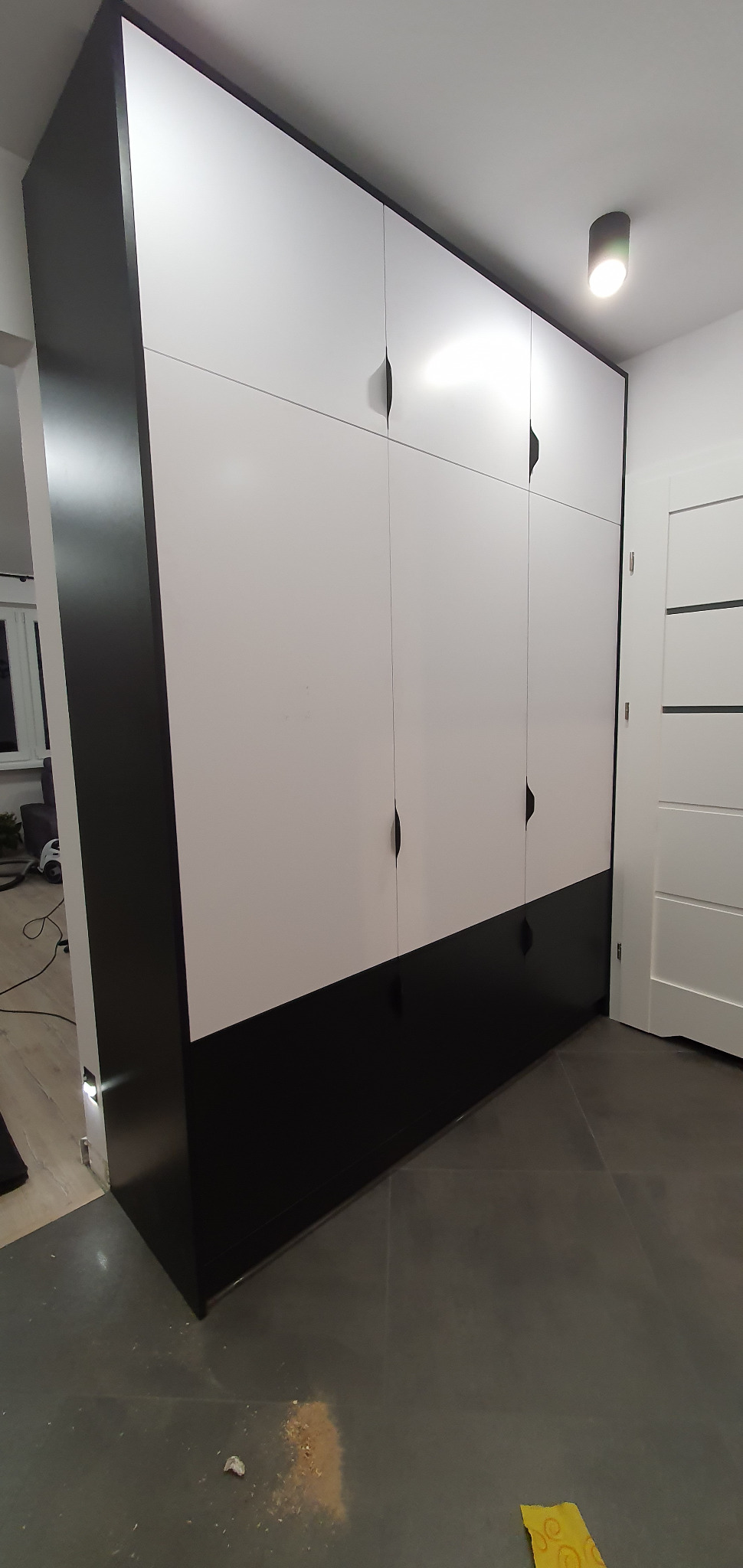 Nowoczesna szafa z białymi frontami i czarnym dołem oraz bokami, z minimalistycznymi uchwytami, stojąca na szarej podłodze, widoczne ślady po montażu