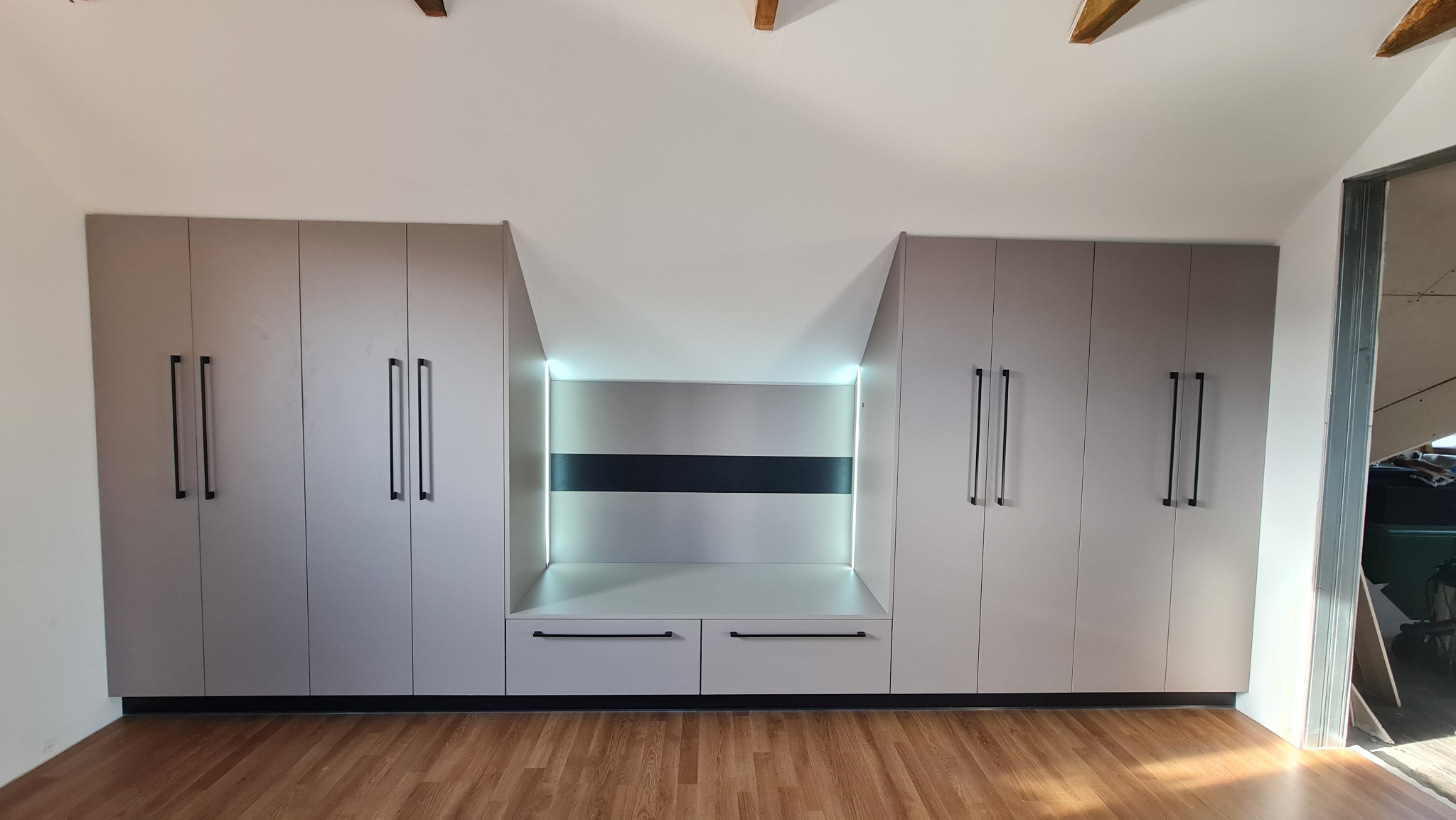 Jasnoszara zabudowa meblowa pod skosem z szafami po bokach i podświetlaną wnęką z szufladami na środku, czarne, minimalistyczne uchwyty.