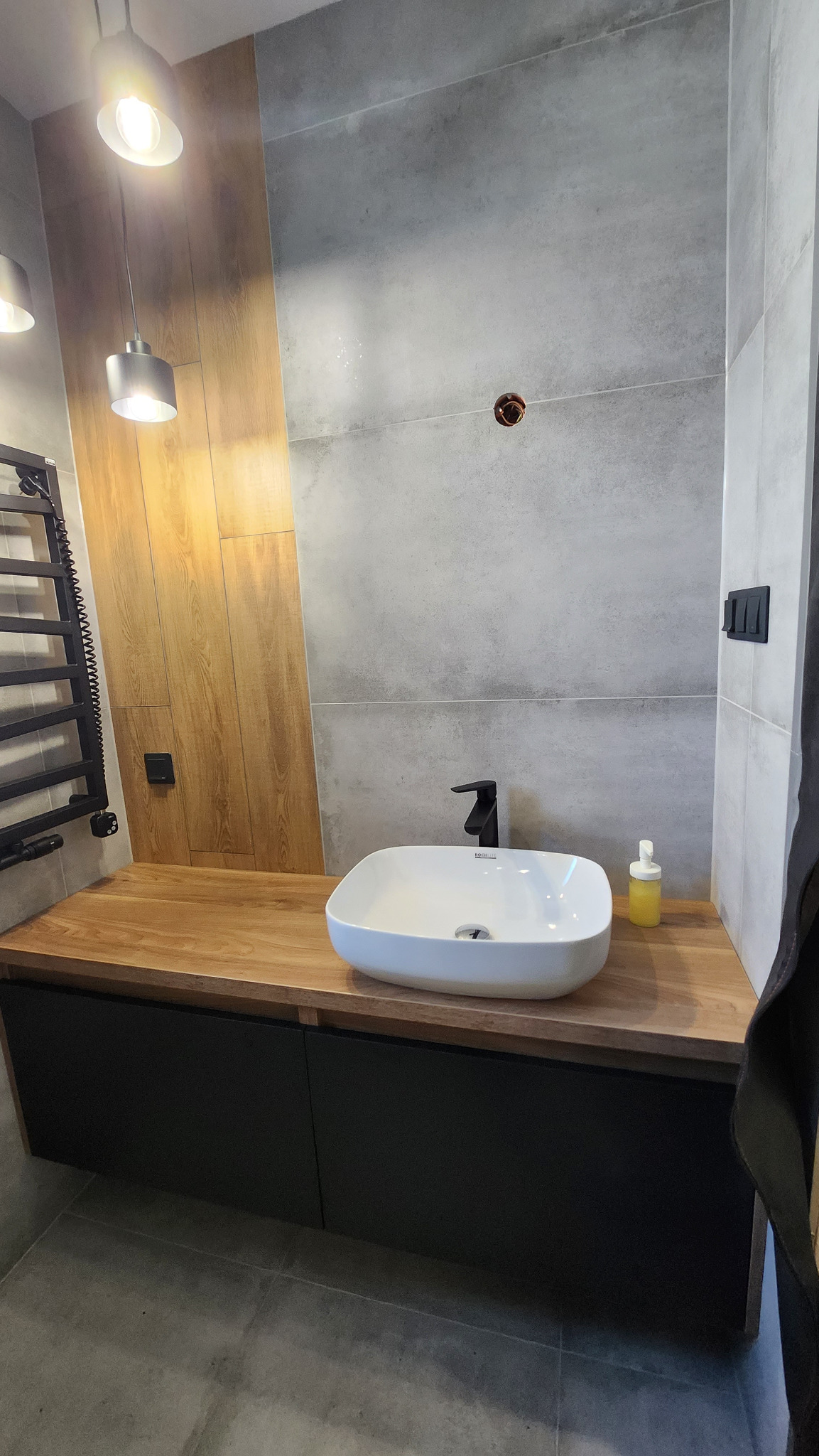 Nowoczesna łazienka z umywalką nablatową na drewnianym blacie i czarnymi szafkami, szare płytki na ścianach, oświetlenie punktowe.