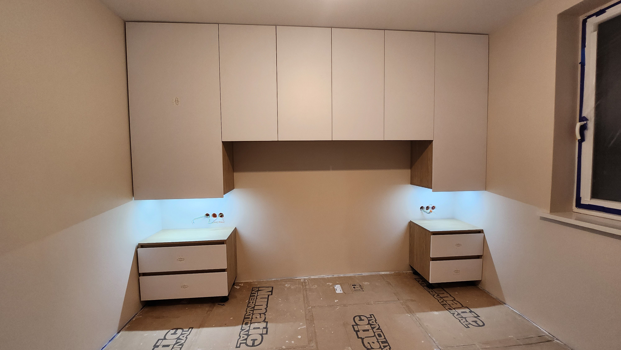 Białe szafki wiszące nad nocnymi stolikami z podświetleniem LED, widoczne kable elektryczne, podłoga zabezpieczona kartonem.