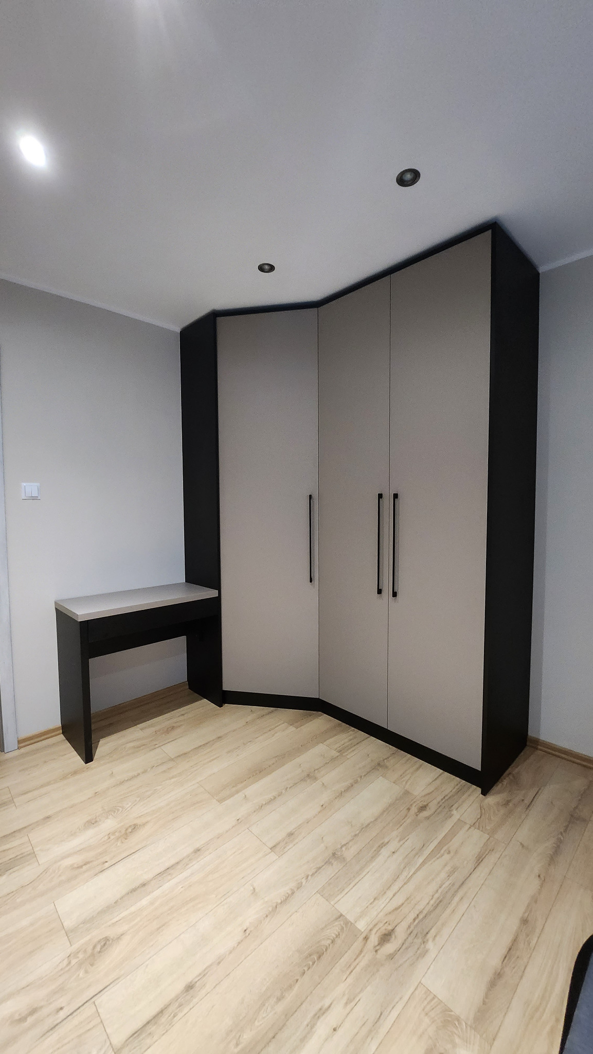 Narożna szafa z frontami w odcieniach szarości i czarnymi detalami, dopasowana do niej toaletka w tym samym stylu, na podłodze panele imitujące drewno.