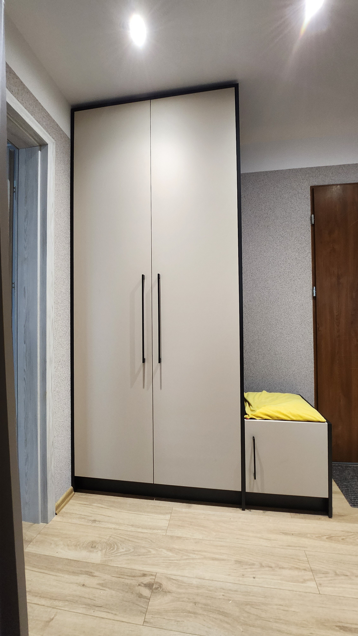 Dwudrzwiowa szafa z jasnymi frontami i czarnymi obramowaniami oraz podłużnymi uchwytami, obok szafki z żółtym materiałem na górze, na drewnianej podłodze w pomieszczeniu z szarą tapetą.