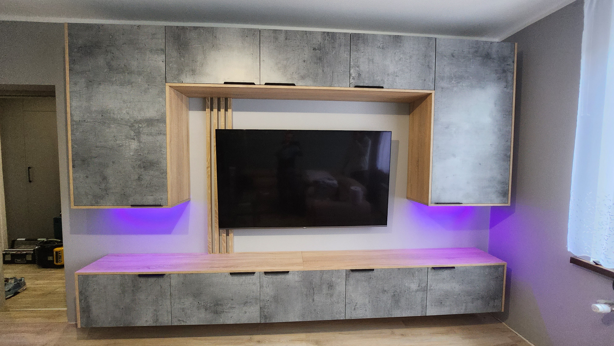Szara szafka RTV na wymiar z podświetleniem LED w kolorze fioletowym, z górnymi szafkami i drewnianymi pionowymi elementami dekoracyjnymi wokół telewizora.